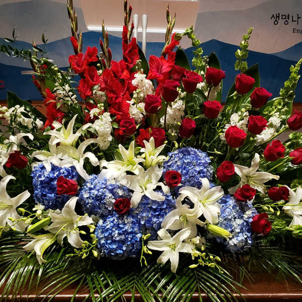 Large red, white, and blue altar arrangement with roses, lilies, hydrangeas, and tall gladiolus.