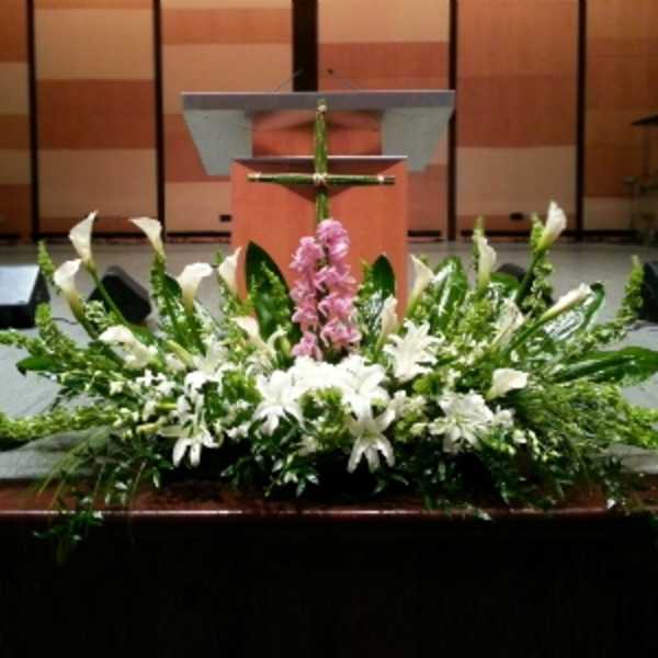 Large white and green altar arrangement with lilies, calla lilies, and a pink flower cluster beneath a green cross.