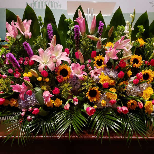 Large horizontal event arrangement of pink lilies, tulips, sunflowers, and mixed blooms with bold tropical foliage