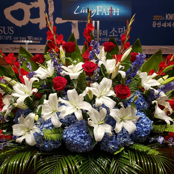 Large red, white, and blue stage arrangement with lilies, roses, hydrangeas, and palm fronds.