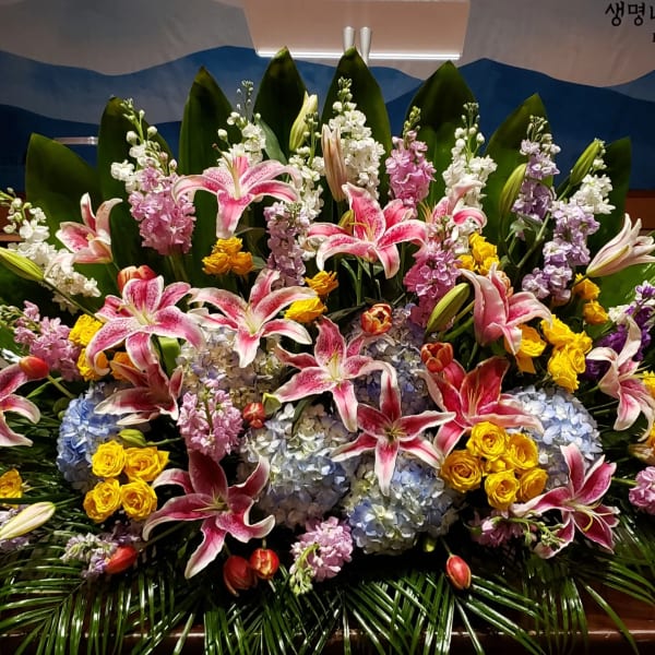 Large fan-shaped arrangement of pink lilies, yellow roses, blue hydrangeas and pastel blooms framed by glossy green leaves
