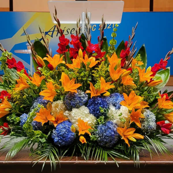 Large altar floral arrangement with orange lilies, blue and white hydrangeas, and red accent flowers on a stage