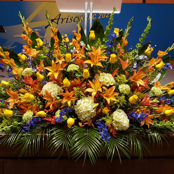 Large altar arrangement with orange lilies, yellow roses, blue delphinium, and white hydrangeas in a fan shape
