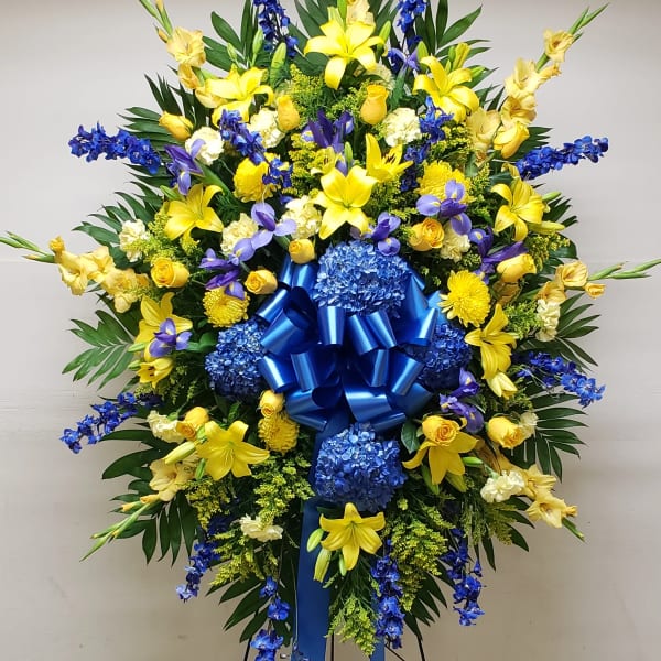 Large standing floral spray with yellow lilies, blue flowers, and a blue ribbon