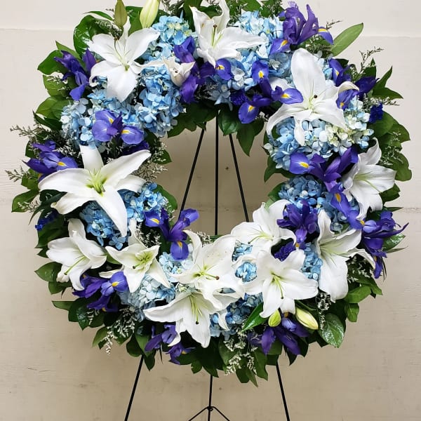 Standing open wreath of white lilies, blue hydrangeas, and purple iris on a metal easel.