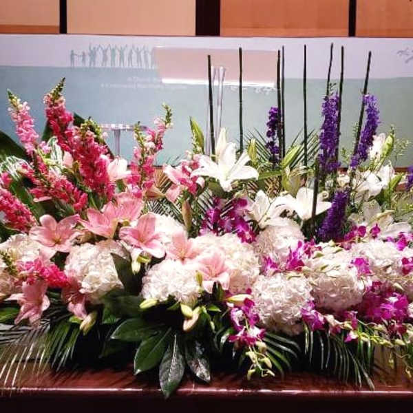 Large low centerpiece of pink lilies, white hydrangeas, and purple flowers along a stage edge