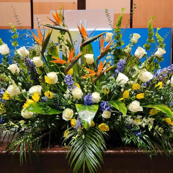 Large stage arrangement of white roses, yellow flowers, blue spikes and orange birds of paradise with lush palm leaves