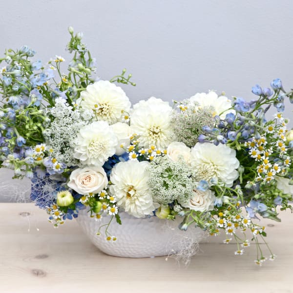 Low white ceramic vase with white dahlias, pale roses, blue delphinium, and small white daisies in an airy arrangement