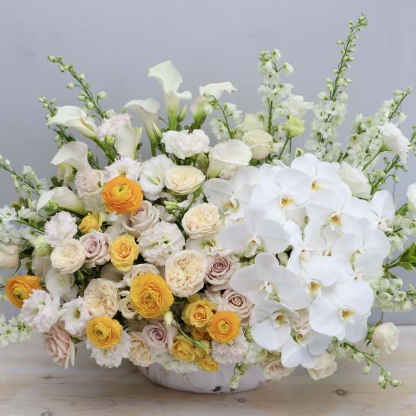 Large low arrangement of white orchids, calla lilies, and yellow and cream roses in a white bowl.