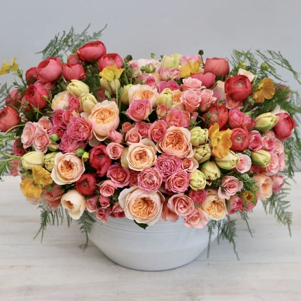 Low white bowl filled with dense pink, peach, and yellow roses and buds in a rounded arrangement.