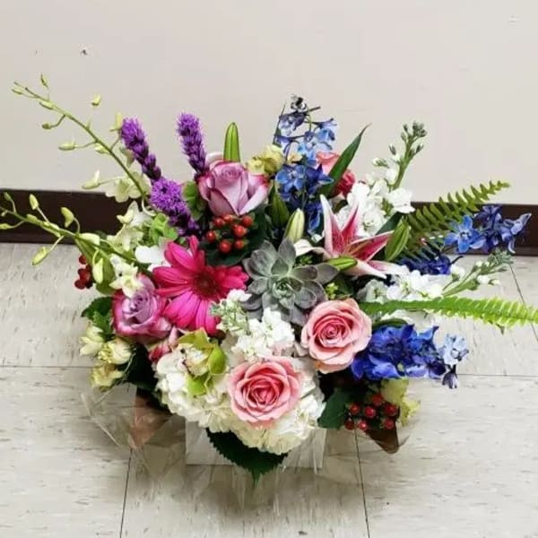 Low mixed arrangement of pink roses, lilies, gerbera daisy, blue delphinium, orchids, and succulents.