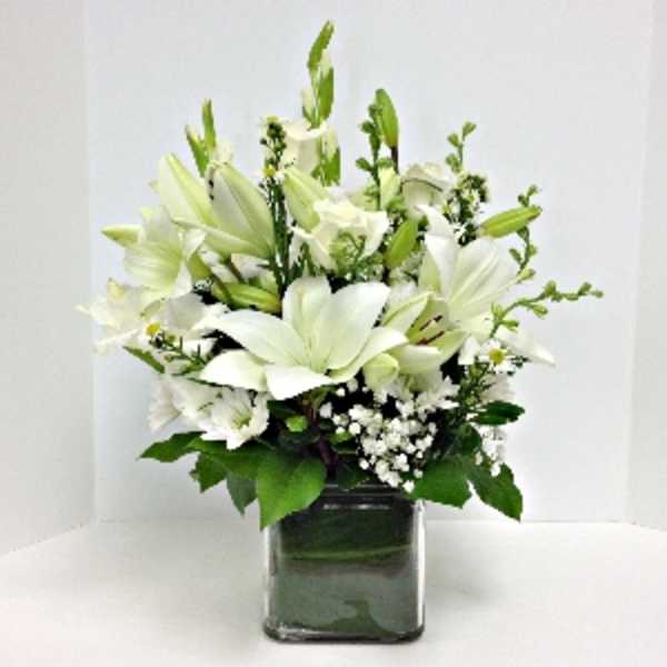 White lilies arranged in a square glass vase with greenery