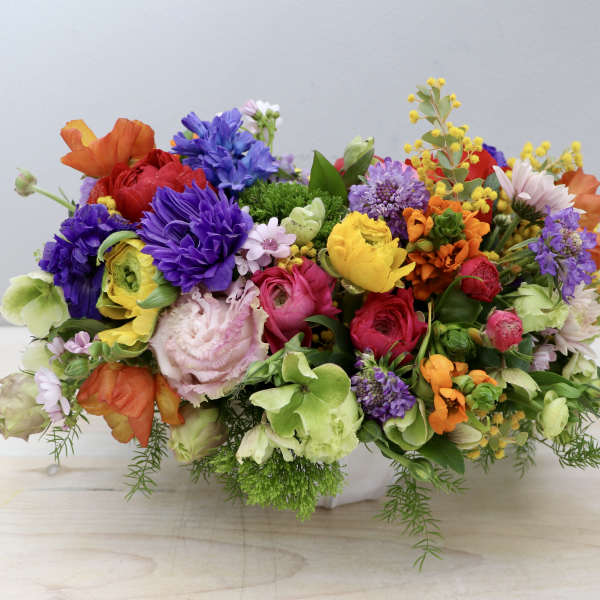 Low, oval arrangement of bright mixed flowers in a white ceramic container on a light wood surface