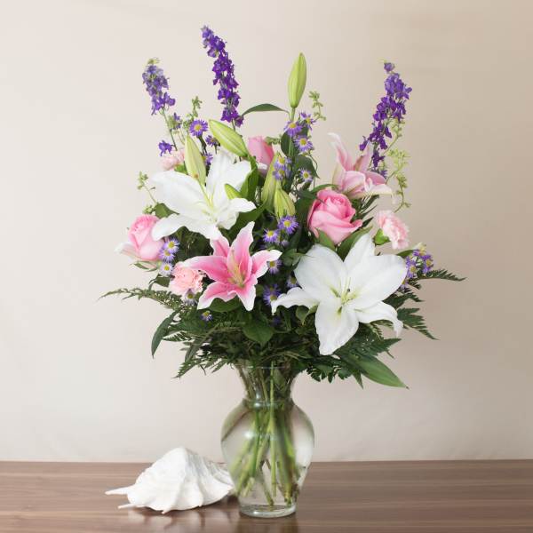 Tall arrangement of white lilies, pink roses, and purple flowers in a clear glass vase.
