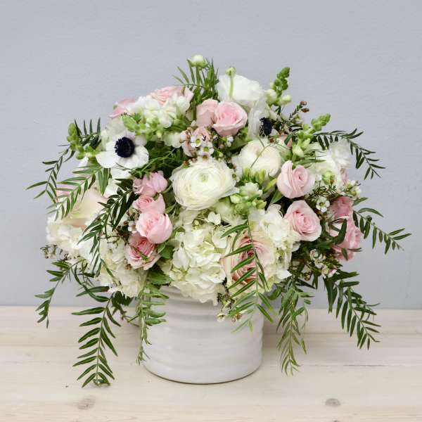White and blush flower arrangement with trailing greenery in a white ceramic vase.