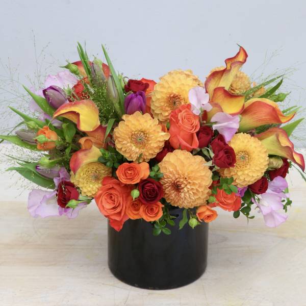Bright mixed arrangement of orange dahlias, roses, and calla lilies in a black cylinder vase