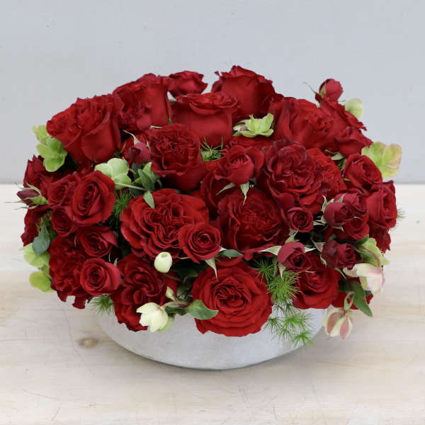 Low round arrangement of red roses in a white ceramic bowl with small green floral accents.