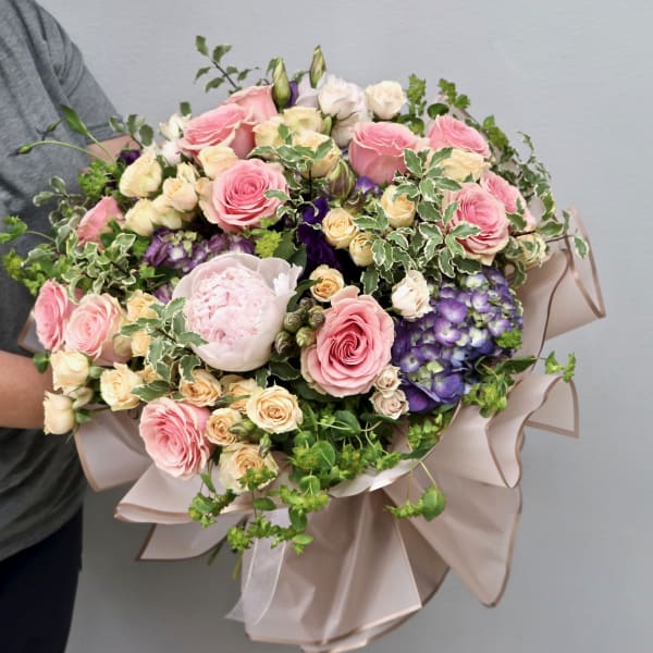 Large hand-tied bouquet of pink roses, cream flowers, and purple hydrangeas wrapped in blush paper