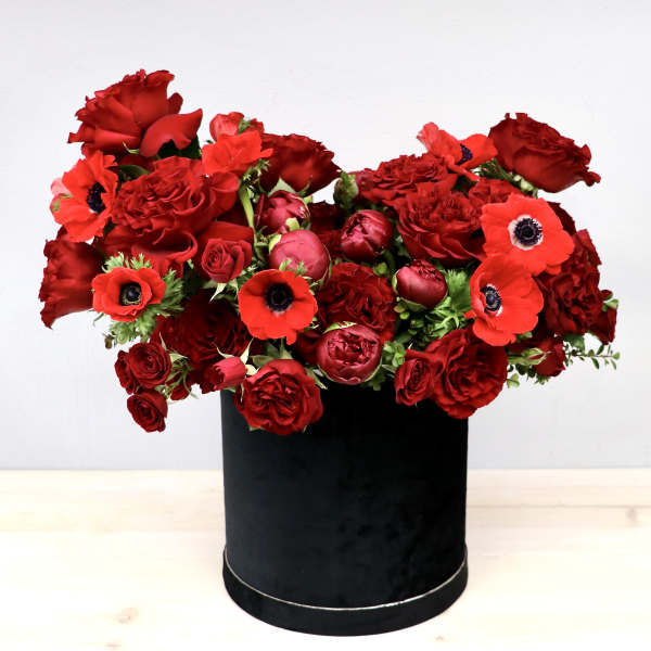 Lush arrangement of red roses, ranunculus, and anemones in a black round hatbox container