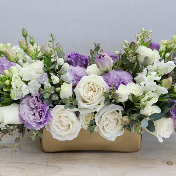 Low arrangement of white roses, purple lisianthus, and white snapdragons in a rectangular gold container.