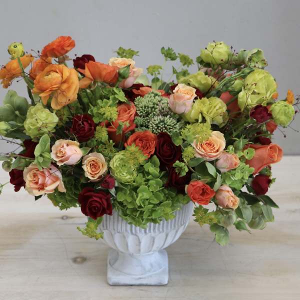 Lush arrangement of orange, peach, red, and green flowers in a white pedestal vase