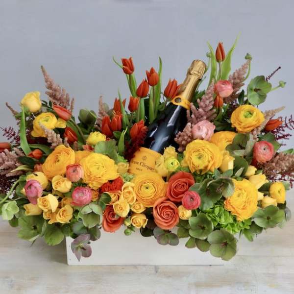 Rectangular arrangement of yellow and orange flowers with red tulips surrounding a bottle of champagne.