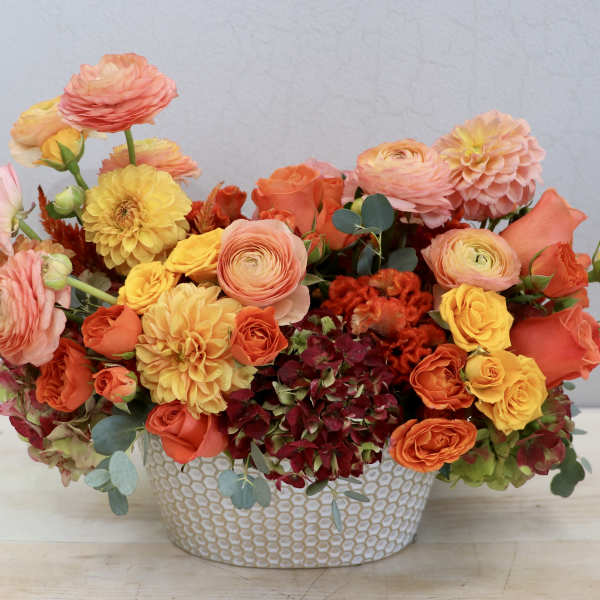 Low arrangement of orange, yellow, and peach flowers in a white patterned ceramic container