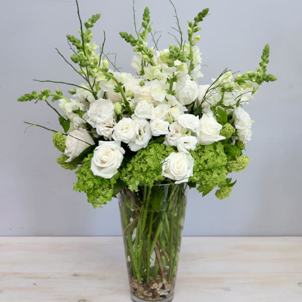 Tall arrangement of white roses and snapdragons with green blooms in a clear glass vase.