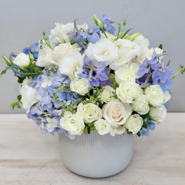 Low white ceramic vase with white roses, blue hydrangeas, and light blue flowers in a rounded arrangement