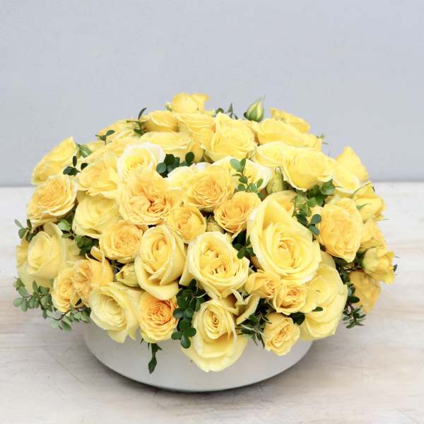 Low round arrangement of yellow roses in a white bowl-shaped container