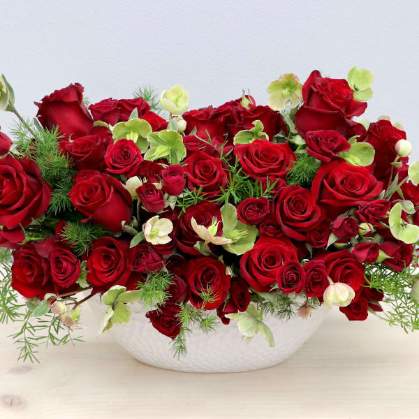 Low oval arrangement of red roses and green blooms in a white bowl.