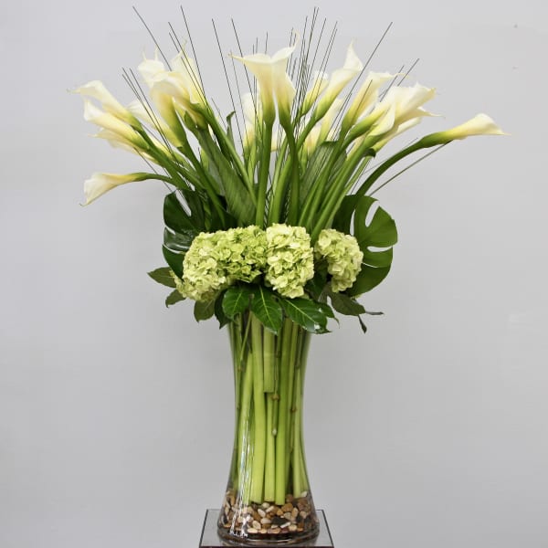 Tall arrangement of white calla lilies and green hydrangeas in a clear glass vase with pebbles