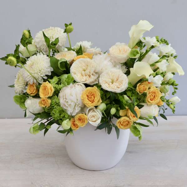 Low arrangement of white and yellow roses, dahlias, and calla lilies in a round white ceramic pot