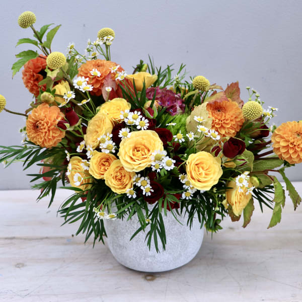 Low arrangement of yellow roses, orange dahlias, and small white daisies in a white pot