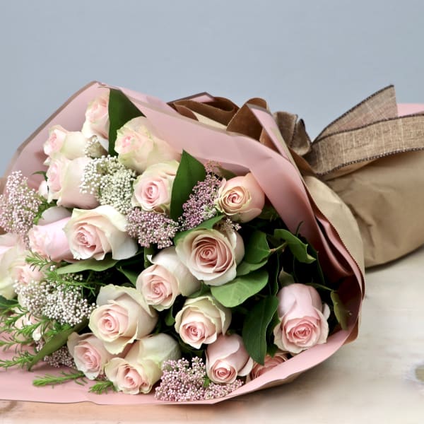 Hand-tied bouquet of pale pink roses with white filler flowers wrapped in pink and kraft paper
