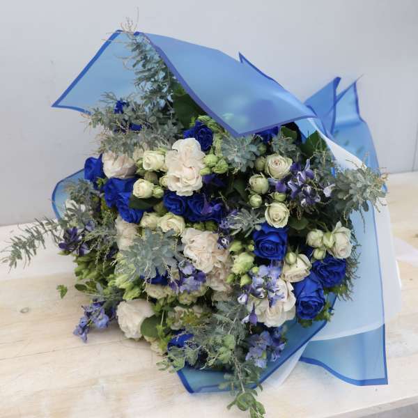 Bouquet of blue and cream roses with small white buds wrapped in blue cellophane.