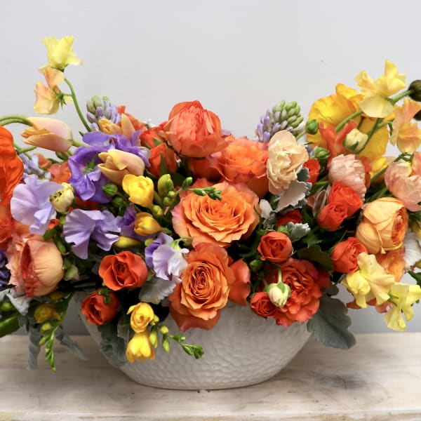 Low ceramic bowl filled with orange roses, ranunculus, and mixed yellow and lavender blooms in a lush arrangement