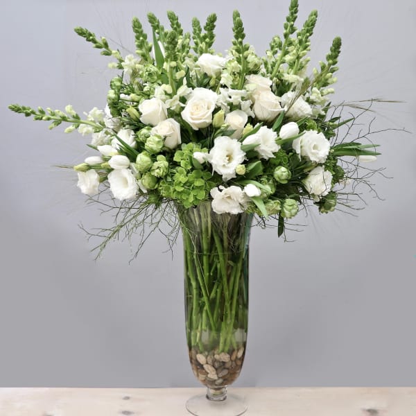 Tall white and green floral arrangement with roses and snapdragons in a clear glass vase with stones
