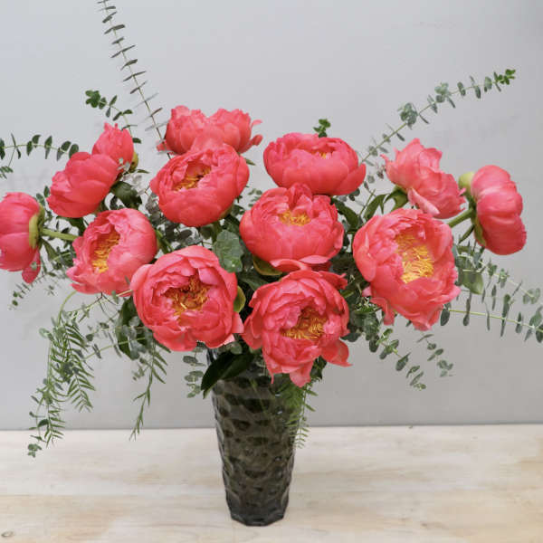 Tall arrangement of bright pink peonies with trailing eucalyptus in a textured glass vase