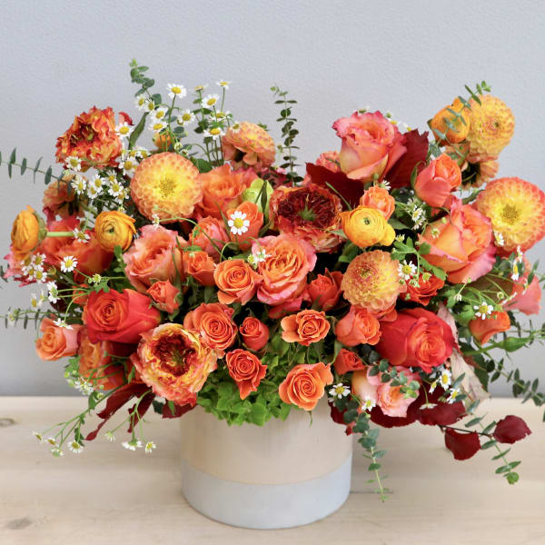 Low arrangement of orange and yellow roses and dahlias in a round white ceramic vase