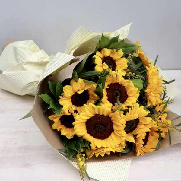 Wrapped bouquet of bright yellow sunflowers with brown centers on a light wood surface.