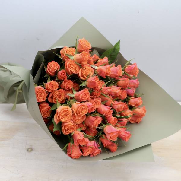 Bouquet of bright orange roses wrapped in matte sage green paper, resting on a light wooden surface