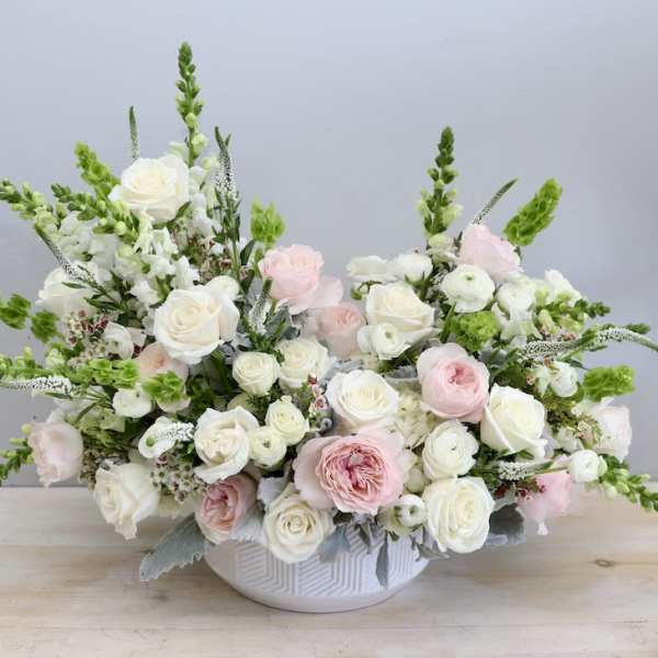 Low white bowl of white and blush roses with green accents in a wide, airy arrangement