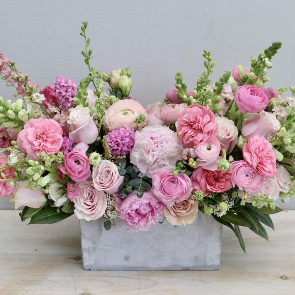 Low rectangular centerpiece of pink roses, ranunculus, peonies and snapdragons in a concrete container.