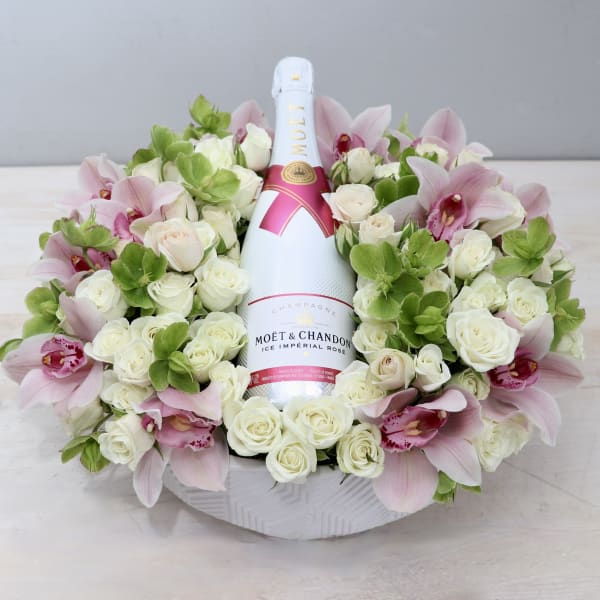 Bottle of champagne nestled in a low ring of white roses, green blooms, and pink orchids in a white bowl