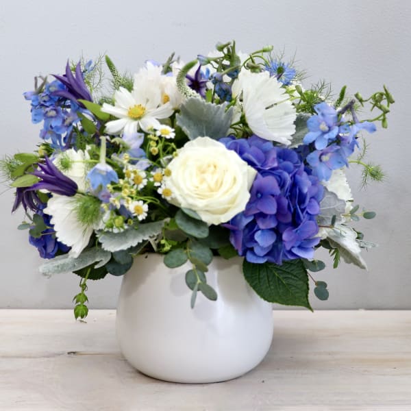Short blue and white flower arrangement with hydrangeas and roses in a round white vase