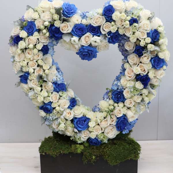 Heart-shaped standing wreath of blue and ivory roses on a moss-covered rectangular base.