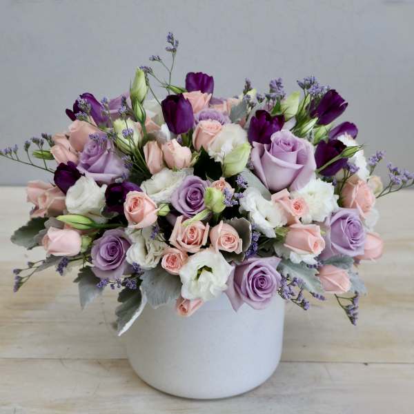 Low pastel arrangement of pink and lavender roses with white blooms in a white ceramic pot