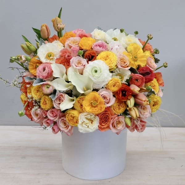 Round arrangement of pink, yellow, orange, and white flowers in a white hatbox-style container.