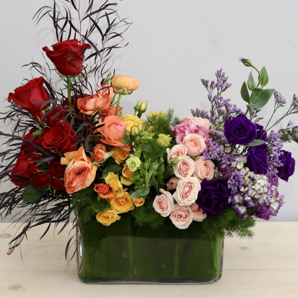 Low rectangular arrangement of red, yellow, peach, and purple flowers in a glass vase.